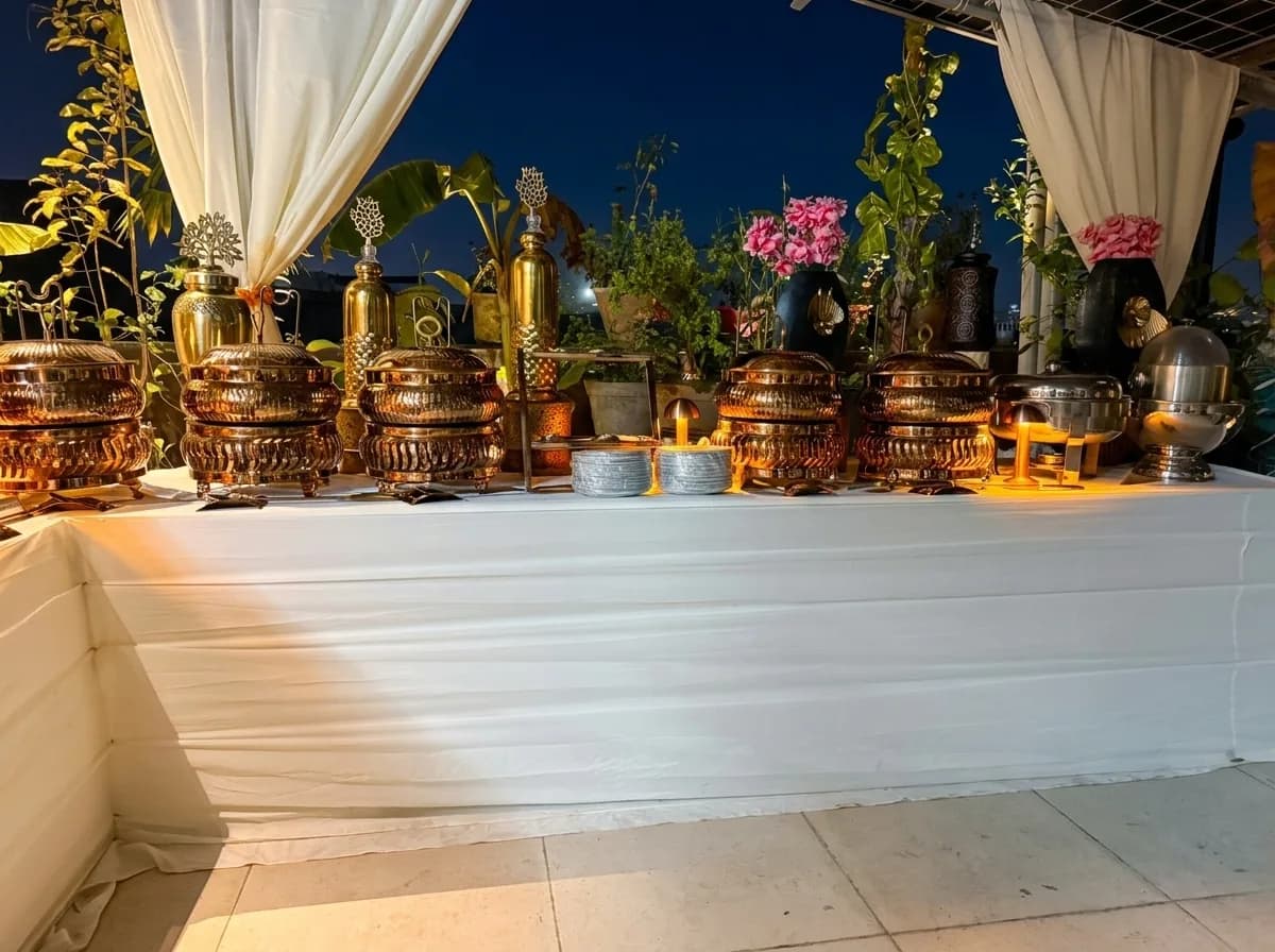 Rooftop terrace buffet at dusk, brass chafing dishes lined along a draped counter under string-lit greenery